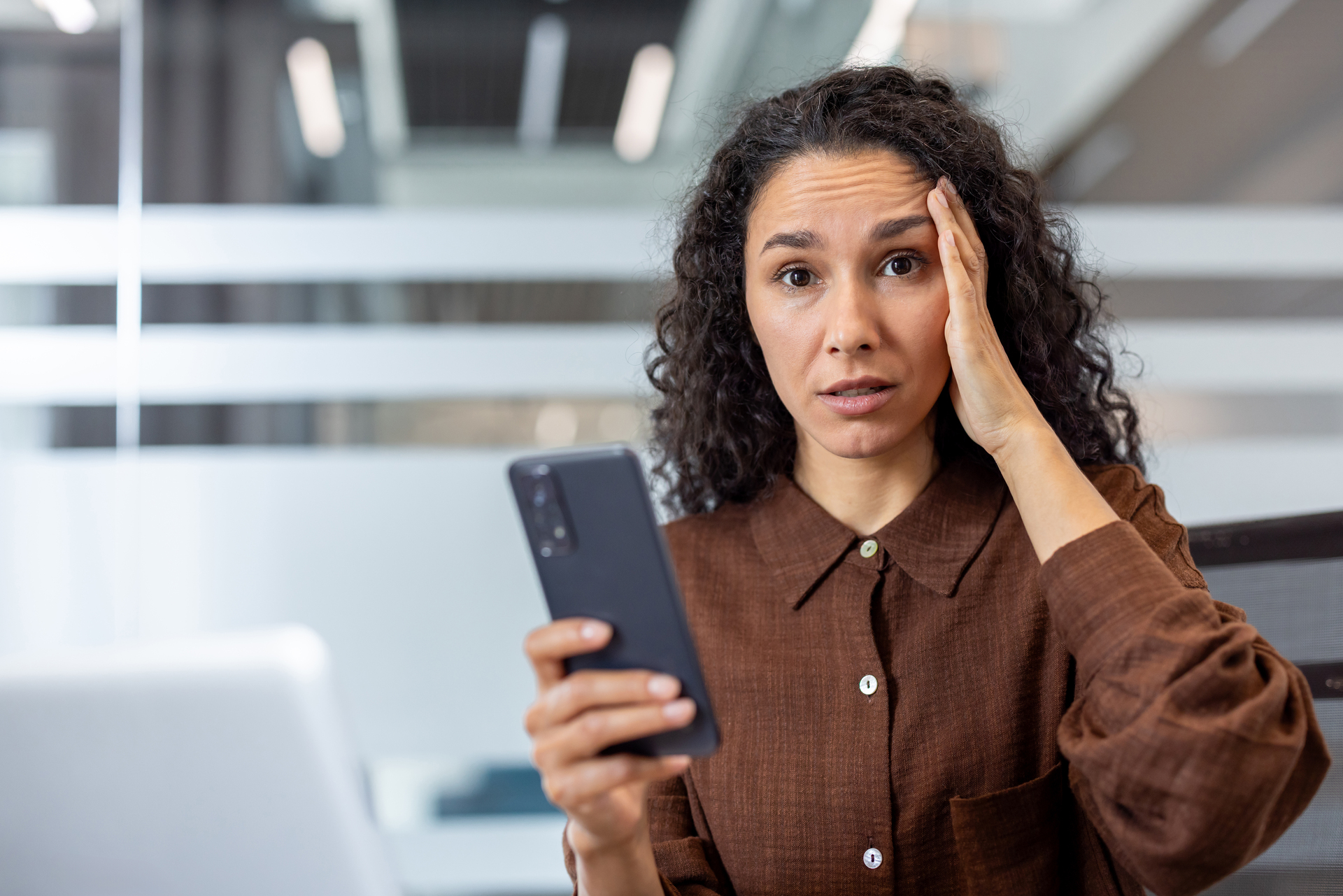 A concerned businesswoman holds her phone, looking stressed with a hand to her head in an office setting.