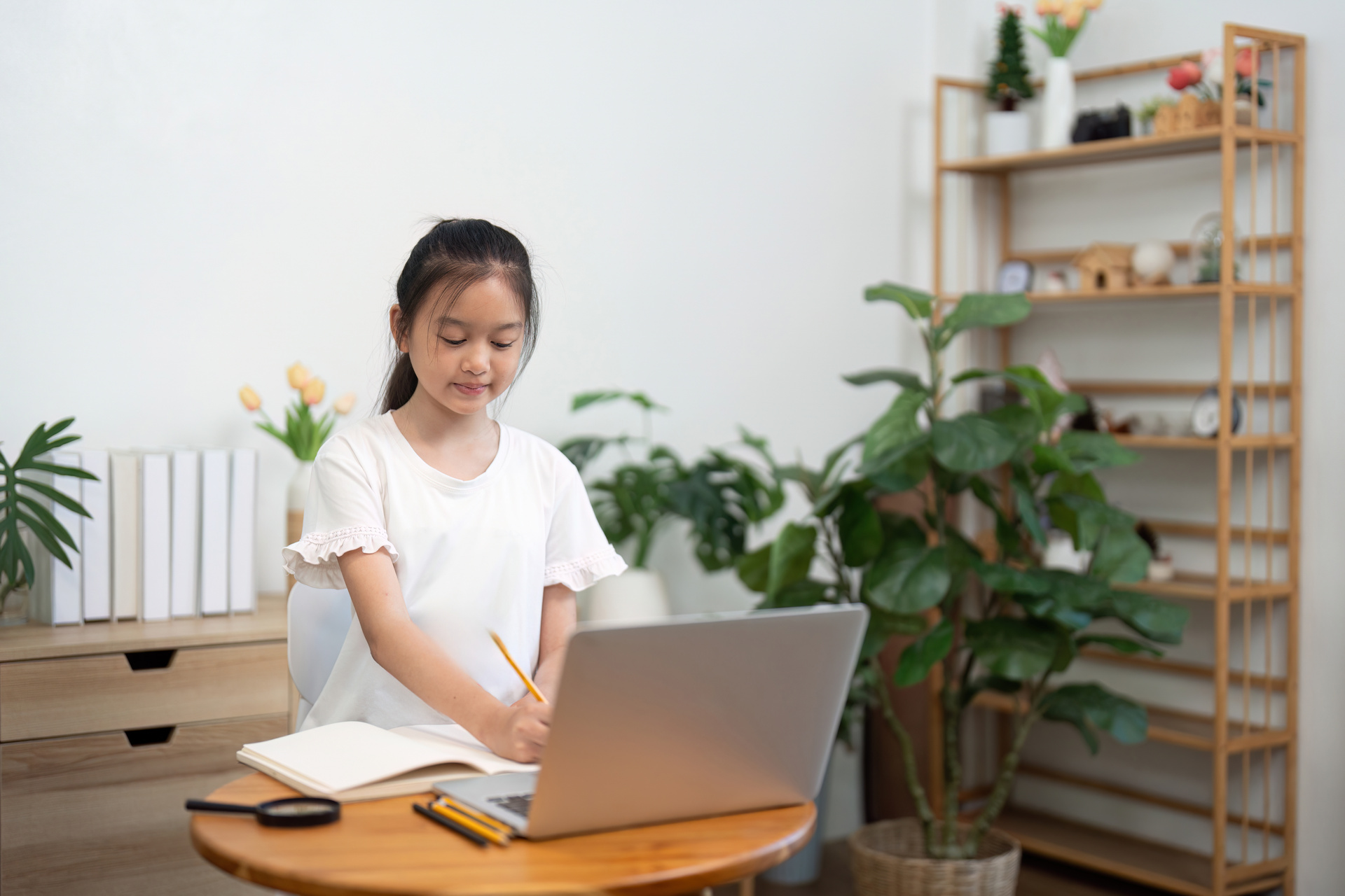 A young girl studying diligently at her desk with a notebook and laptop, working hard in a well-lit room filled with plants.