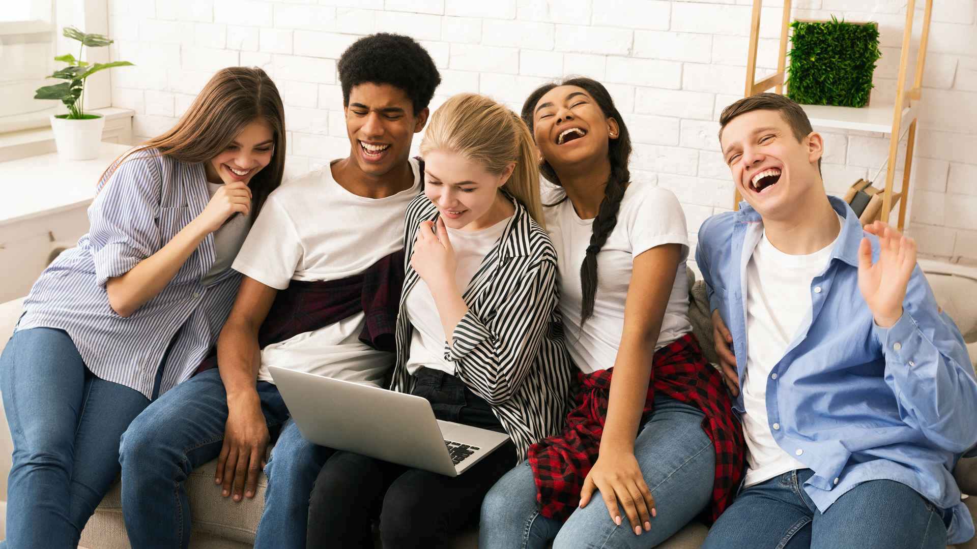Five multiethnic teenagers is gathered cozily on a couch, sharing a joyful moment while looking at a laptop screen. Their expressions range from broad smiles to laughter